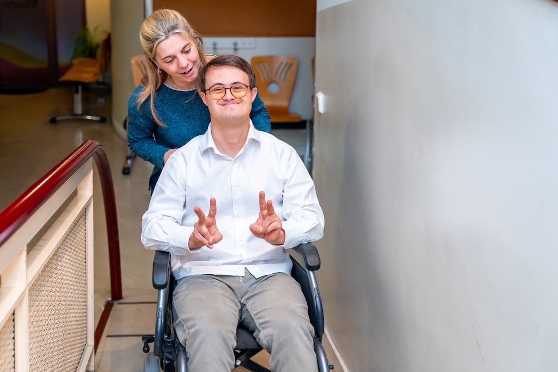 Disability support worker pushing a happy man with down syndrome using wheelchair while he gesturing success with fingers